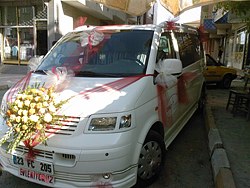 Beyaz minibüsün önünde sarı çiçekli süsleme ve kırmızı kurdele
